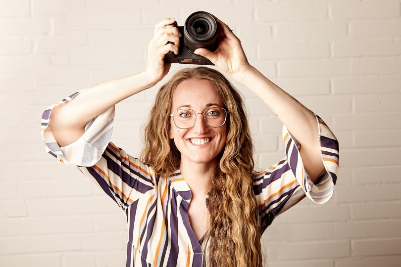 Die Fotografenmeisterin Sonja Schröder aus Lehrte hät ihre Kamera in beiden Händen über den Kopf und lächelt in die Kamera. Sie steht vor einer weißen Ziegelwand und trägt ein blau-weiß-gelb gestreiftes Hemd.